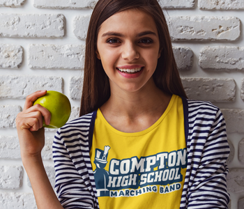 A girl creatively showcasing an apple while posing against a brick wall in her eye-catching homecoming shirt design.